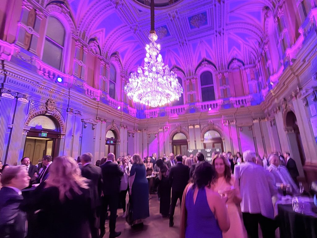 A large group of people in formal attire mingle in an ornate hall with high ceilings, arched windows, and a chandelier, under purple lighting—capturing the excitement of the Sydney Business Awards Regional Finalist event hosted by Spark Interact.
