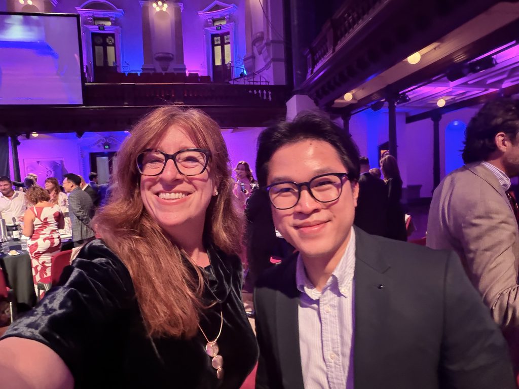 Two people wearing glasses smile for a selfie at an indoor event with purple lighting, likely taken during the Sydney Business Awards, with other attendees and signage for Spark Interact visible in the background.