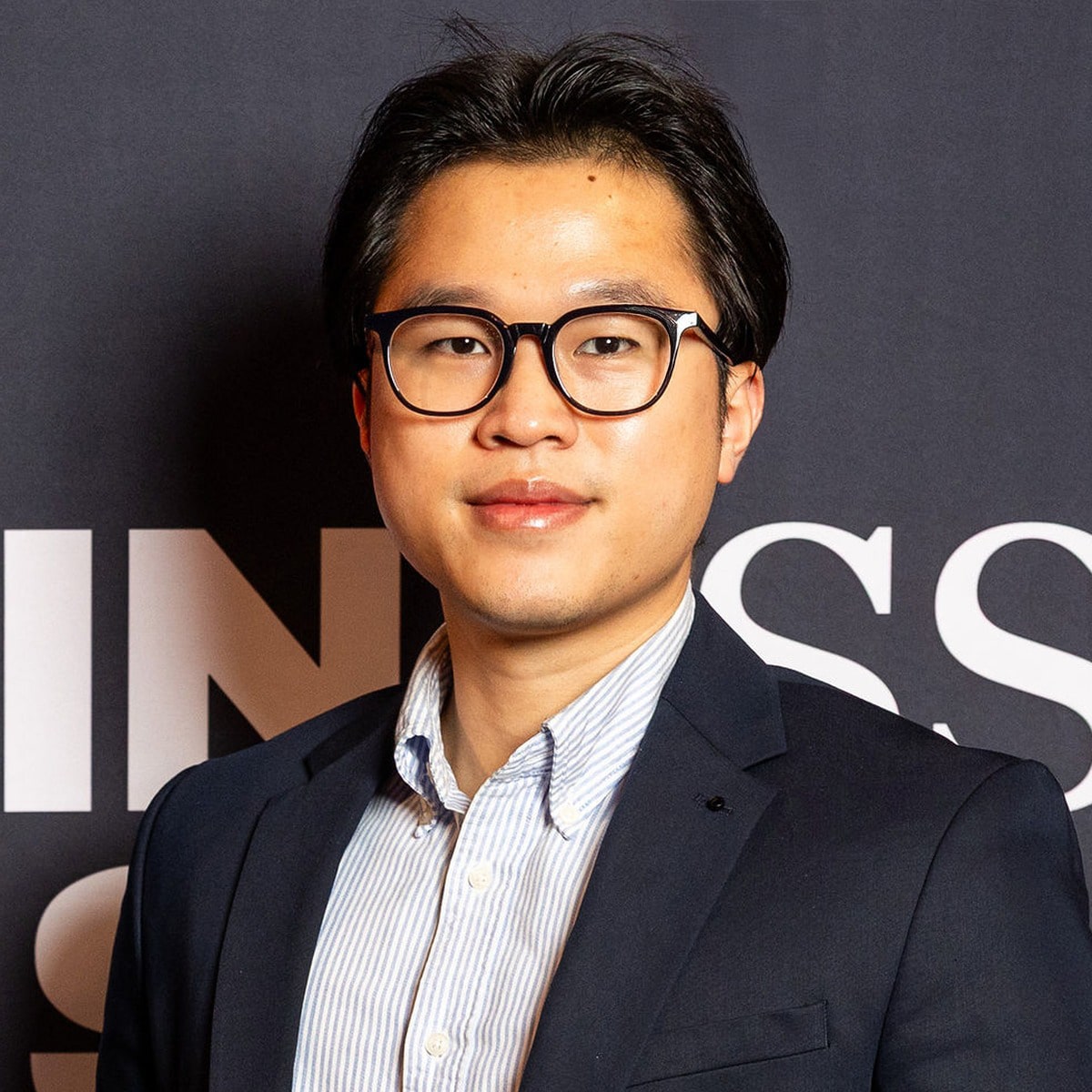 A man with short black hair and glasses, wearing a navy blazer and striped shirt, stands in front of a black background with partial white text, presenting a professional image suited for the business and AI industries.