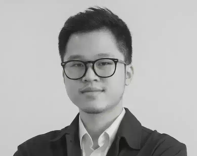 A man wearing glasses, a buttoned shirt, and a jacket stands with arms crossed against a plain background.